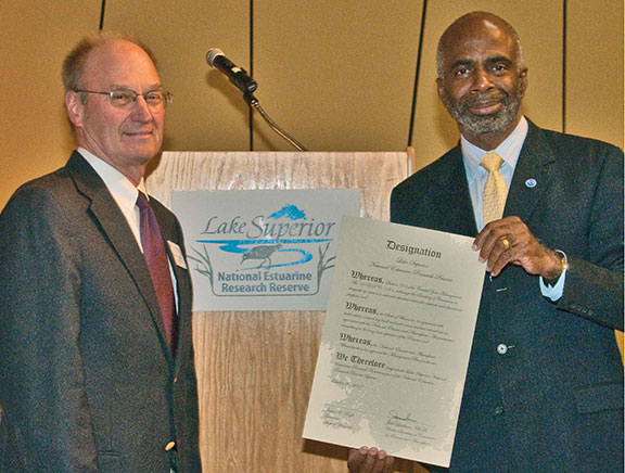 Two men standing in front of a podium. One man is holding an official designation document.