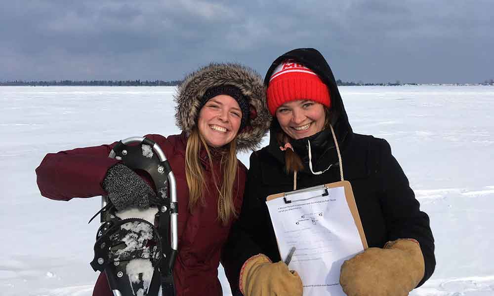 Two young women wearing winter clothes, smiling and holding a snow shoe and a clipboard. They are standing on a vast area of snow covered lake ice.