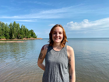 A girl with red hair wearing a gray tank top standing in front of a lake