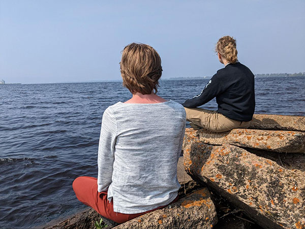 Two women sitting, facing away from the camera, on rocks looking at a lake